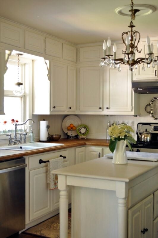 A traditional kitchen with white cabinets, wood countertops, a chandelier, kitchen decorating details like a small island with a vase of flowers, stainless steel appliances, and a window over the sink.