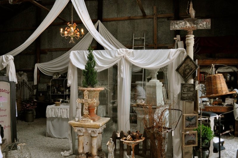 Decorative booth with white drapes and chandelier in a rustic indoor market, displaying vintage items, potted plants, and antiques.
