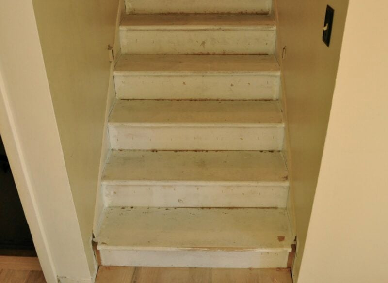 A small stairway with five worn, white-painted wooden steps featuring stained treads and beige walls on either side, leading upward.