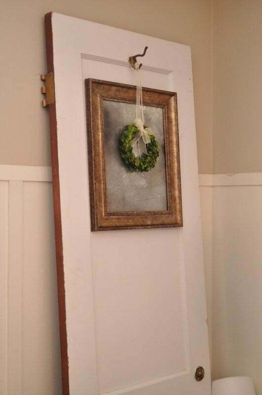 A white wooden door leans against a beige wall, adorned with an antique mirror from a picture frame and a small green wreath hanging on it.