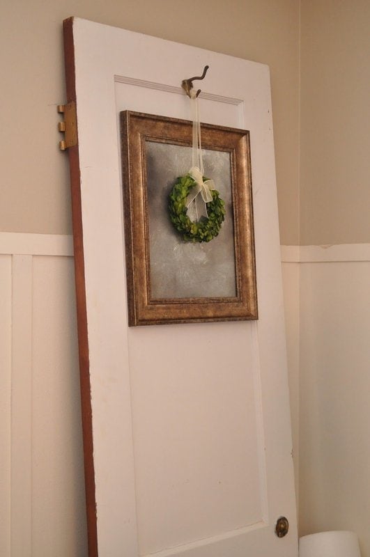 A white wooden door leans against a beige wall, adorned with an antique mirror from a picture frame and a small green wreath hanging on it.