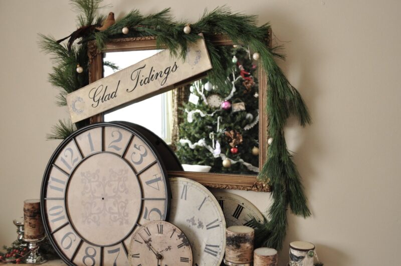 A mirror decorated with pine garland and a "Glad Tidings" sign, with several vintage clocks and candles in front, reflecting a Christmas tree.