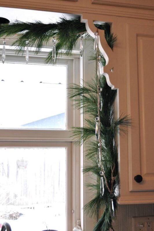 Pine garland and hanging crystal ornaments decorate the corner of a kitchen window with white trim.