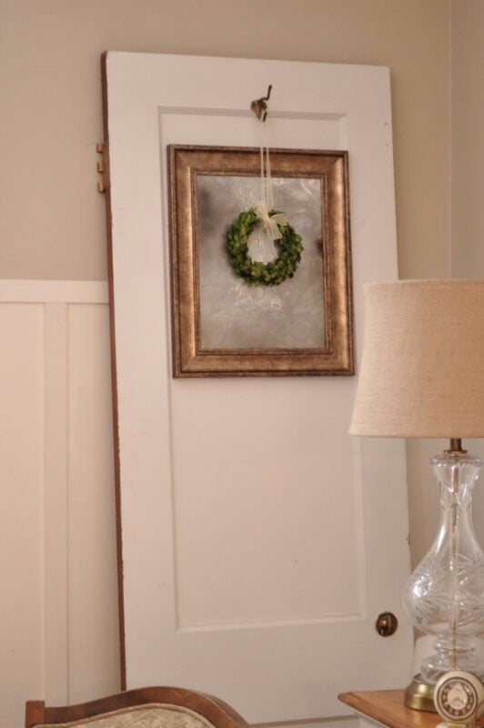 A white door leaning against a beige wall, decorated with a framed wreath, next to a clear glass lamp with a beige shade.