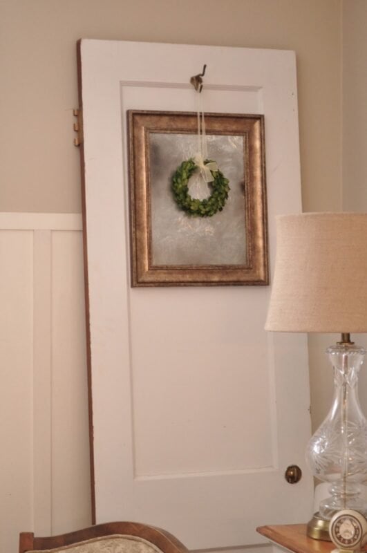 A white door leans against a beige wall, with a framed wreath hanging from a hook, next to an antique mirror made from a picture frame and a clear glass lamp with a beige shade.