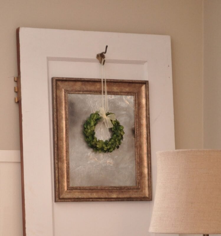 A small green wreath is hung by a ribbon over an antique mirror made from a picture frame on a white door, with part of a beige lampshade visible in the lower right corner.