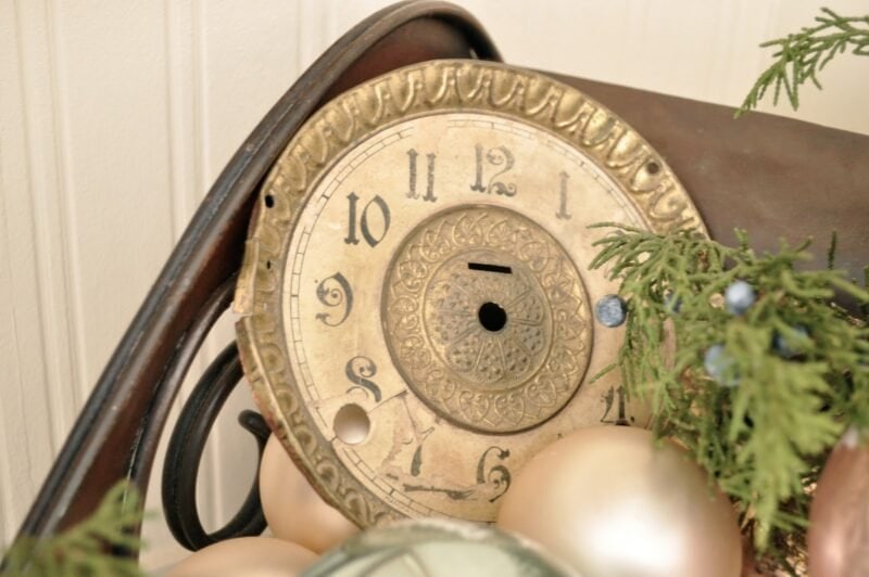 A close-up of a vintage, weathered clock face without hands, leaning on a surface with greenery and ornaments in the foreground.