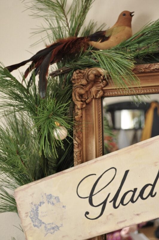 A decorative bird and pine branches sit atop an ornate framed mirror, with part of a sign reading "Glad" visible in the foreground.