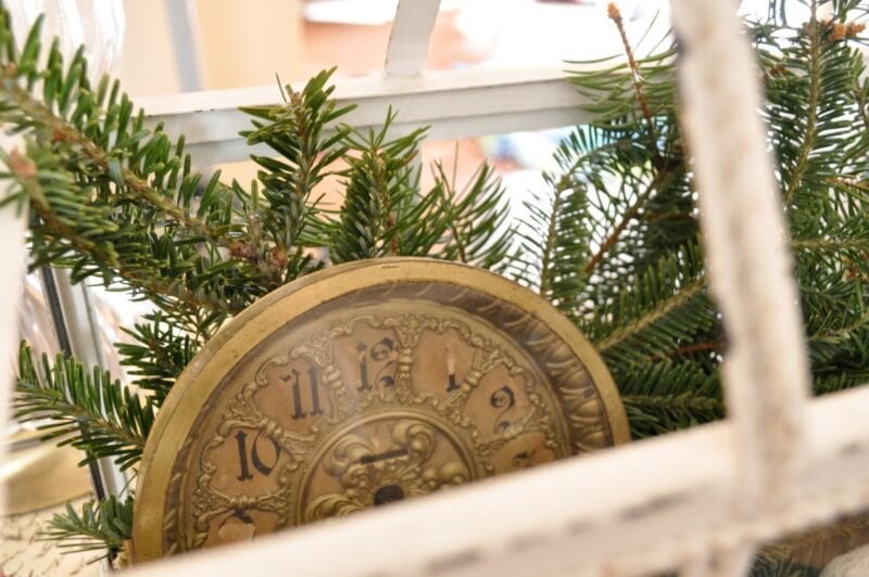 An ornate vintage clock partially covered by evergreen branches, viewed through a white metal frame.