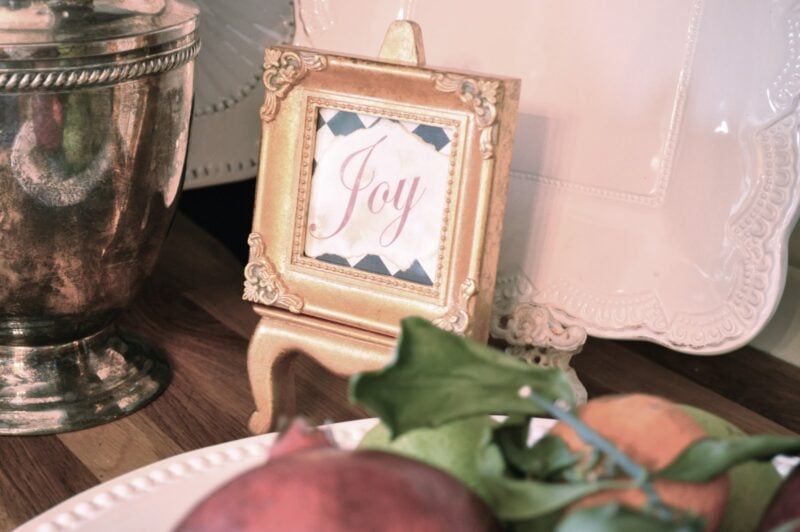 A small gold-framed sign with the word "Joy" sits on a wooden table next to a silver vase and a white plate with fruit.