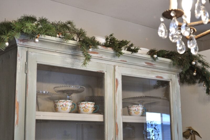 A distressed cabinet with glass doors displays teacups and glassware, topped with a garland of pine branches and ornaments; a chandelier hangs nearby.