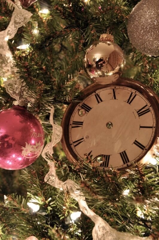 A Christmas tree decorated with a pink ornament, a silver ornament, white lace, and a vintage clock face among the branches.