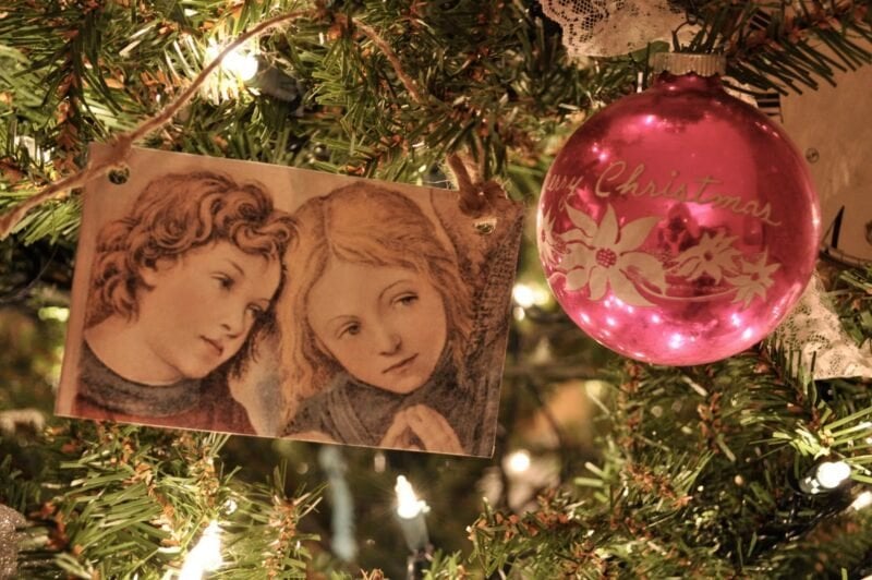 A close-up of a Christmas tree with a Renaissance-style angel illustration and a pink ornament that reads "Merry Christmas.