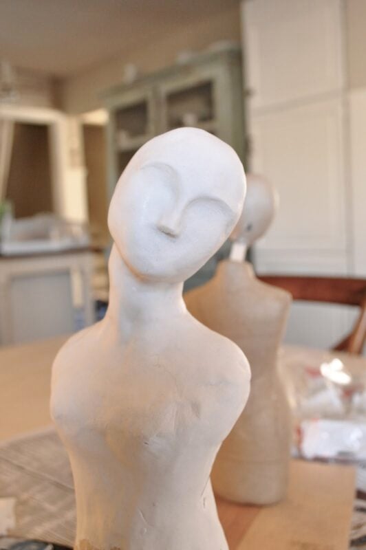 A white, unfinished sculpture of a stylized human bust with a tilted head—reminiscent of sculpting a Santos doll form in clay—sits on a table in a room with blurred furniture in the background.