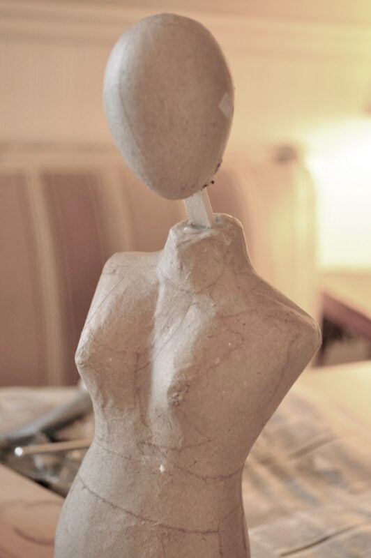 A papier-mâché mannequin torso with an oval head, reminiscent of sculpting a Santos doll form in clay, features no facial details against a neutral background.