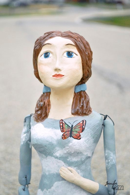 A painted wooden Sculpted Santos Art Dolls sculpture of a girl with brown hair in pigtails, blue eyes, and a butterfly on her chest stands on a paved surface.
