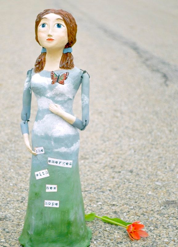 A sculpted Santos Art Doll of a woman with braided hair and a butterfly on her dress stands on a road beside a fallen red flower; words on her dress read "she emerged with new hope.