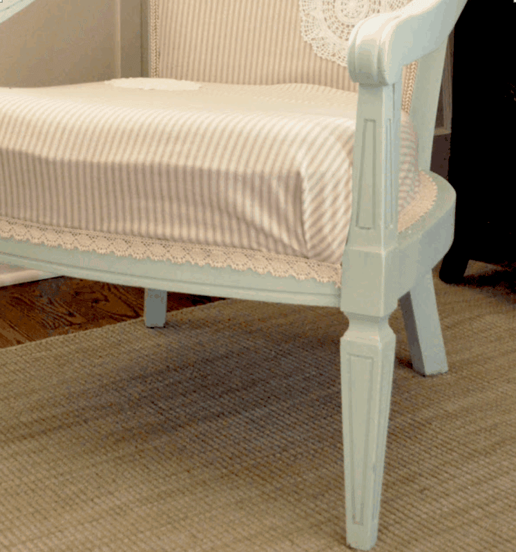 Light blue wooden chair with beige and white striped cushion and lace trim, placed on a hardwood floor next to a wall—perfect inspiration for embellishing a chair with doilies for added vintage charm.