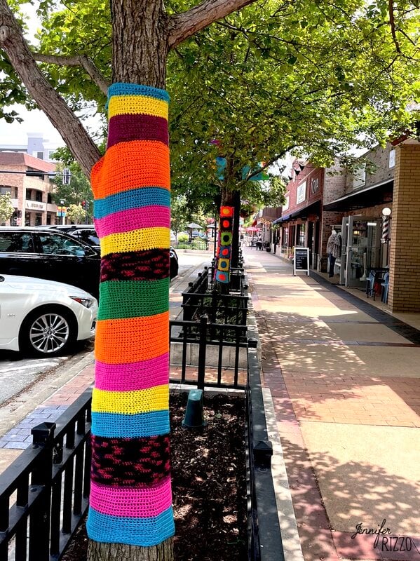 A tree trunk on a city sidewalk is wrapped in colorful crochet yarn, with more crochet wrapped yarn trees lining the street in vibrant patterns.