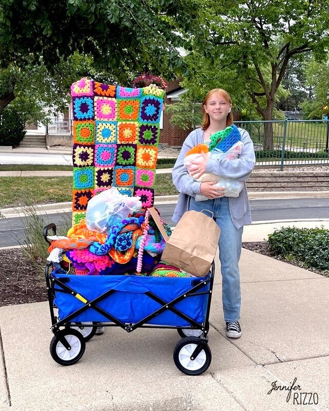 A young person stands outdoors beside a blue wagon filled with colorful crocheted blankets and a brown paper bag, holding more crochet items in their arms, near crochet wrapped yarn trees.