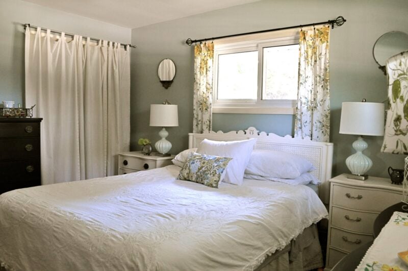 A neatly made bed with white linens is centered between two nightstands with lamps, under a window with floral curtains in a softly lit bedroom.