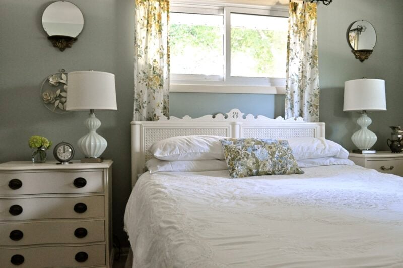A neatly made bed with white bedding and floral pillow is between two nightstands with lamps, set against a wall with a window and floral curtains.