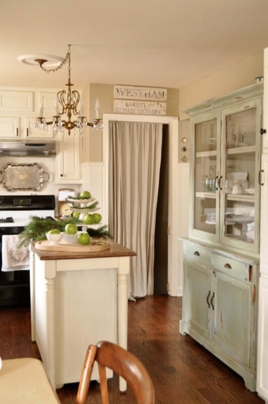 A kitchen with white cabinets, a chandelier, a small island with green apples, a vintage hutch, and a curtain covering a doorway.