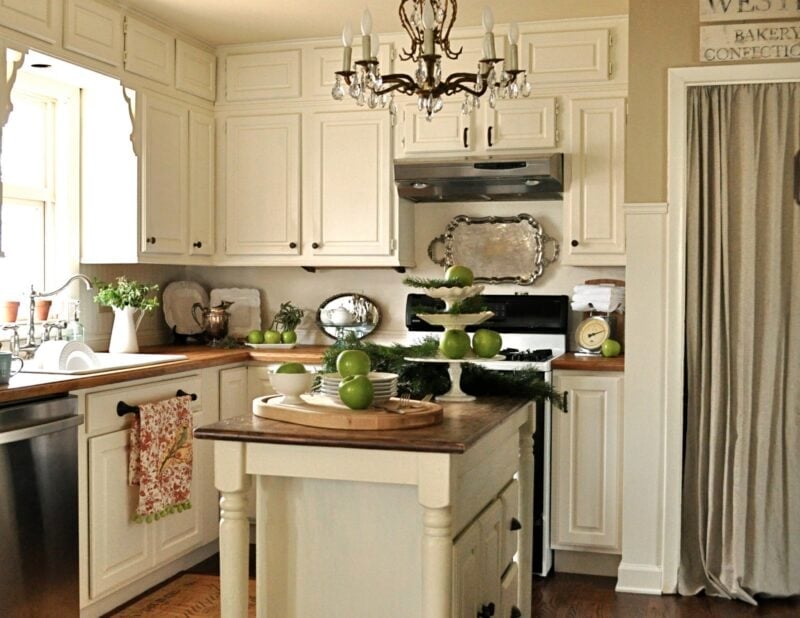 A kitchen with cream-colored cabinets, a central island with green apples, stainless steel appliances, a chandelier, and a curtain covering a pantry area.