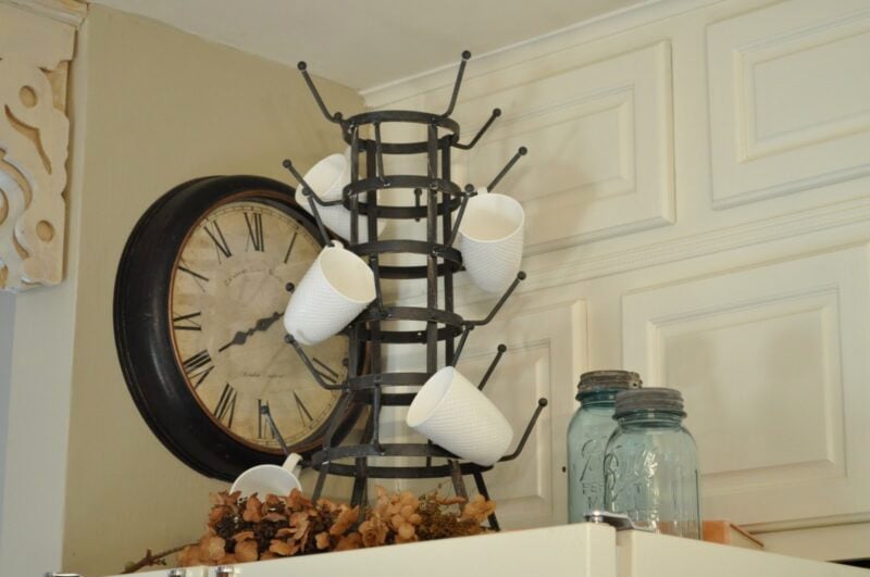 A metal mug rack holds several white mugs on top of a cabinet, next to a large wall clock and two blue mason jars.