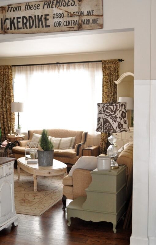 A cozy living room with beige armchairs, a cream sofa, a round wooden coffee table with a potted plant, and patterned curtains framing a large window.