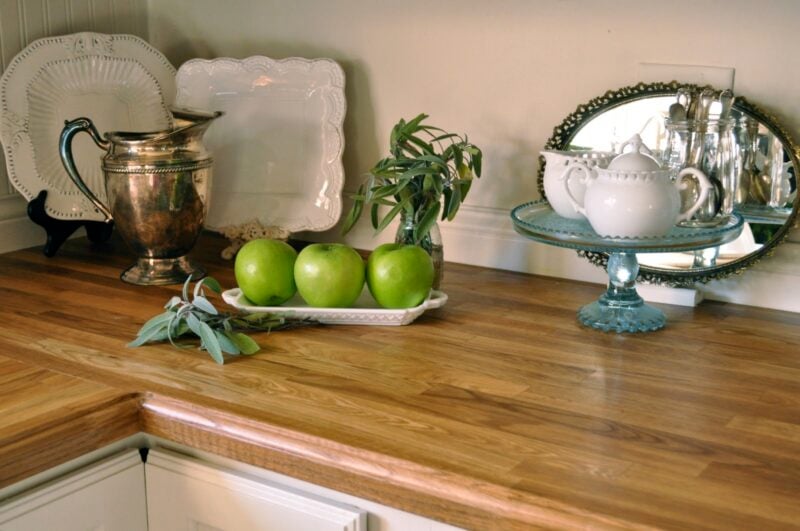 A wooden countertop displays three green apples on a white tray, silverware, a teapot on a glass stand, and decorative plates, with a small mirror in the background.