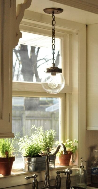 A glass pendant light hangs above a kitchen sink, with potted herbs on the windowsill and sunlight streaming through the window.