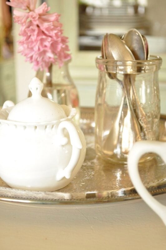 A white sugar bowl, a glass jar with spoons, and a small vase with a pink flower are arranged on a silver tray.