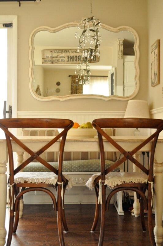 A dining area with a white table, two wooden chairs, a bench, a lamp, a bowl of fruit, and a wall mirror reflecting part of the room and chandelier.