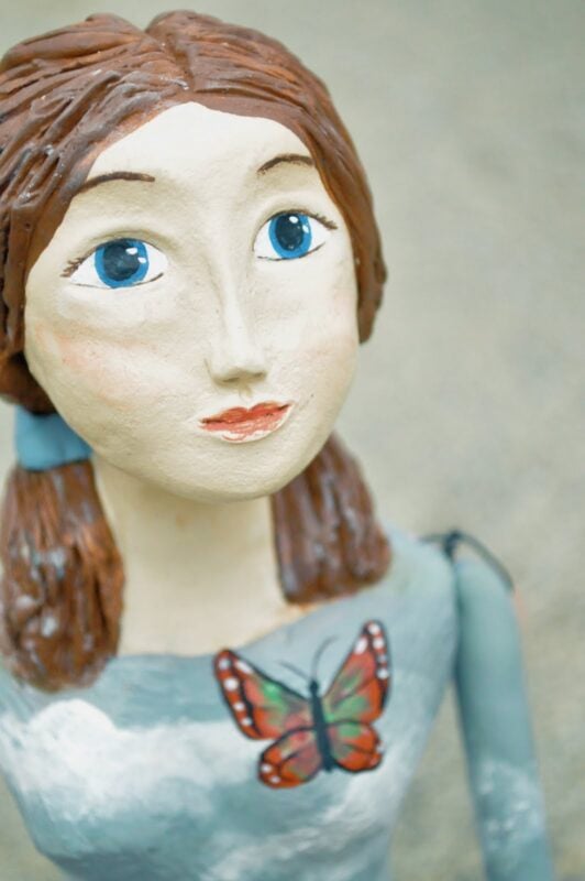 A close-up of a painted clay figurine with blue eyes, brown hair, and a butterfly design on its shirt, reminiscent of The Urban Journey Upcycled Clothing and Vintage Collection on Etsy, set against a plain background.