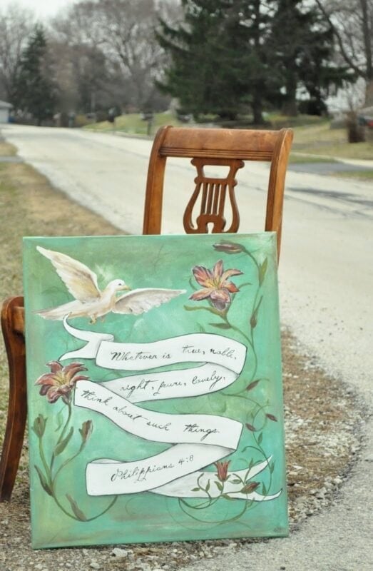 A wooden chair sits by a roadside, beside a painted canvas featuring a dove, flowers, and Philippians 4:8 on green—an inspiring scene reminiscent of The Urban Journey Upcycled Clothing and Vintage Collection on Etsy.