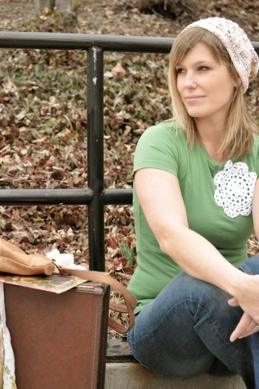 A woman wearing a green t-shirt and jeans from The Urban Journey Upcycled Clothing and Vintage Collection on Etsy sits outdoors on a concrete step near a suitcase and dry autumn leaves, looking to the side.