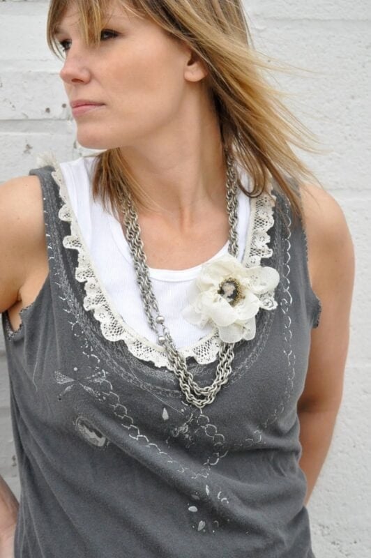 Woman wearing a grey sleeveless top with a lace-trimmed neckline, layered over a white shirt and styled with a large flower necklace, stands against a white wall—discover similar looks in The Urban Journey Upcycled Clothing and Vintage Collection on Etsy.
