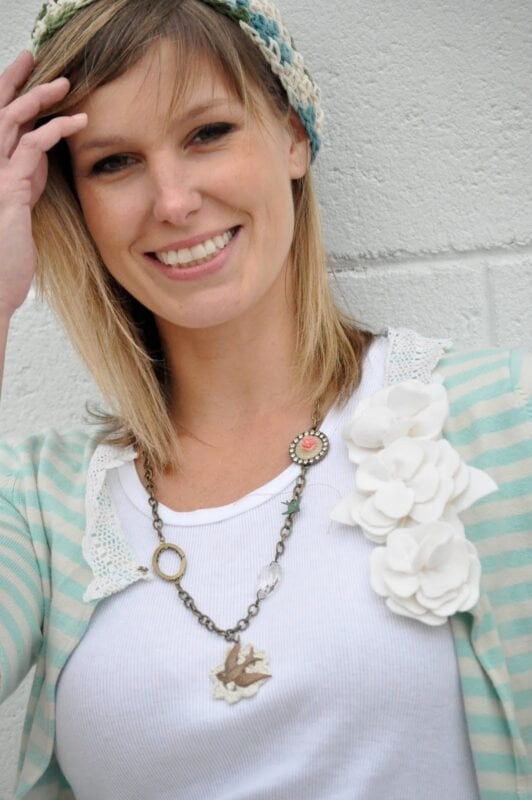 Woman smiling in a striped cardigan and white shirt with fabric flower embellishments from The Urban Journey Upcycled Clothing and Vintage Collection on Etsy, accessorized with a beaded necklace and crochet hat, standing against a textured wall.