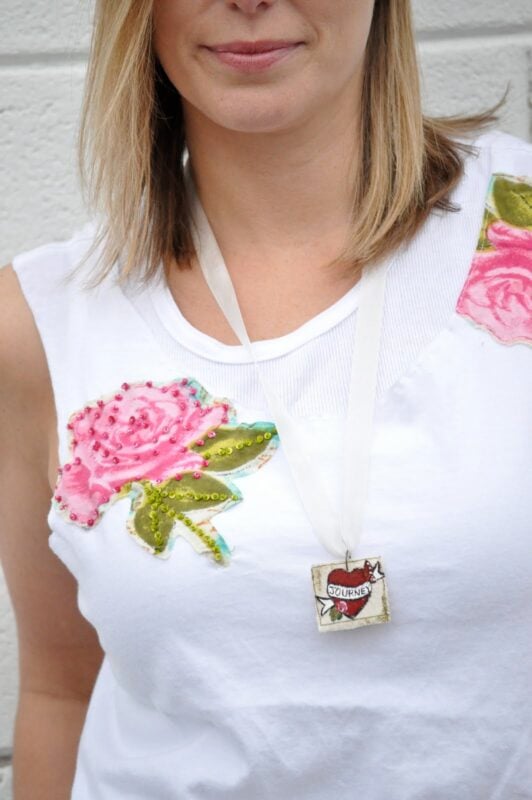 A woman wearing a white sleeveless shirt with pink flower appliqués and a square pendant necklace from The Urban Journey Upcycled Clothing and Vintage Collection on Etsy, featuring a bird and the word "journey.