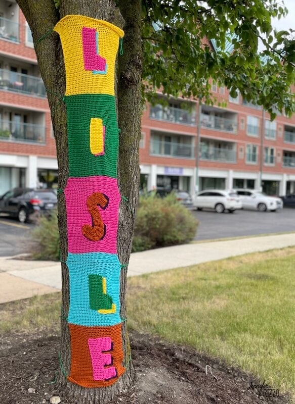 A tree trunk is crochet wrapped in colorful knitted panels, each displaying a large letter that together spell "LOSSLE." An apartment building and parked cars are visible in the background.