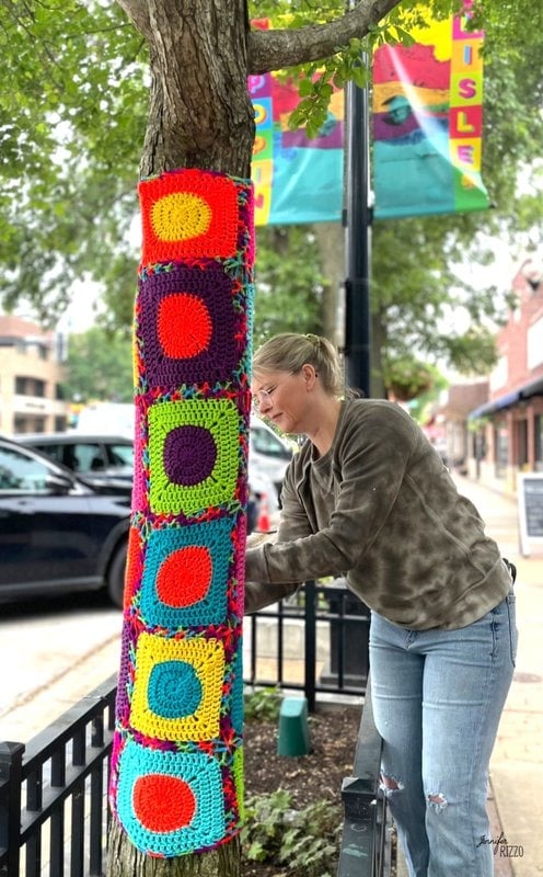 A woman decorates a tree trunk with crochet wrapped yarn squares on a city sidewalk, bright banners fluttering and parked cars in the background.