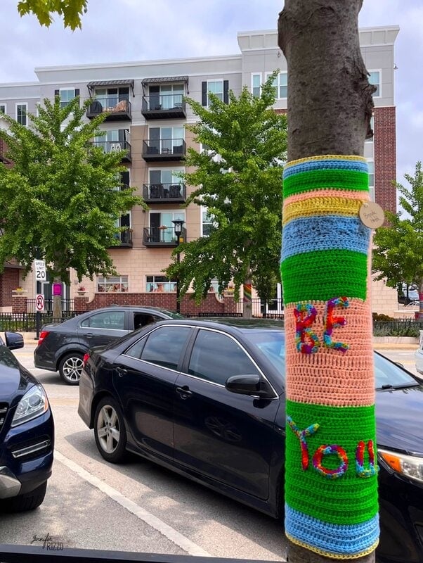 A crochet wrapped yarn tree stands on a city street, its vibrant colors spelling out "BE YOU." Cars line the road and an apartment building rises in the background.