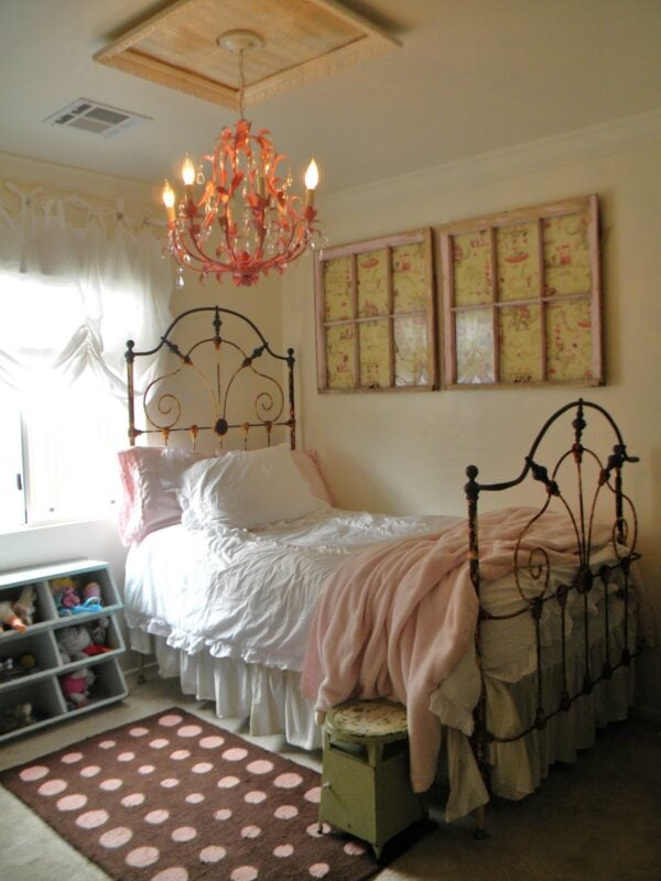A bedroom with a vintage-style metal bed, white and pink bedding, a pink chandelier, wall art, a toy shelf, and a brown rug with pink dots showcases gorgeous decorating ideas for a little girl's bedroom.