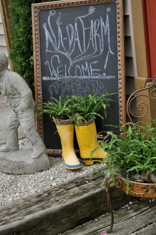 Chalkboard sign reading "Na-Da Farm Welcome" behind yellow boots used as planters with ferns, next to a statue and a potted plant on a wooden step.