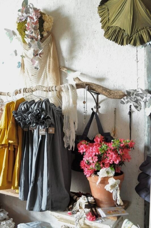 Clothes and scarves hang on a rustic branch rack in this creative retail display; a potted pink azalea sits below, surrounded by fabric and accessories against a textured wall.