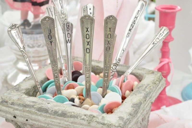 A rustic and creative retail display features a square stone container filled with pastel-colored buttons, holding vintage silverware with words like "XOXO," "JOY," and "LOVE" stamped on the handles.
