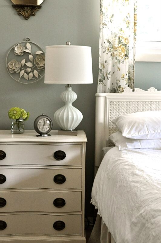 A white nightstand with a lamp, clock, and vase of green flowers sits next to a bed dressed in white linens and floral curtains—what's your style for creating a cozy bedroom retreat?.