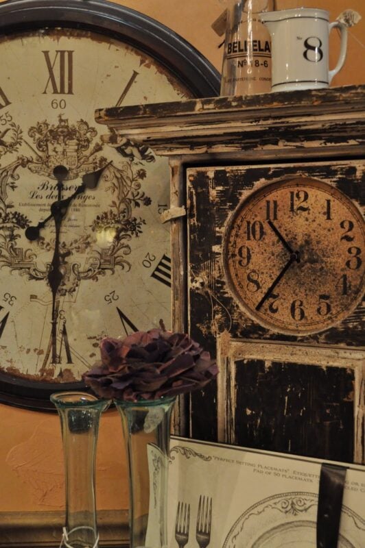 Two antique clocks, one round and one square, add whimsy to Ruby Begonia's Shop retail display, accompanied by glass vases, a mug labeled "8," and a dried flower in front of them.