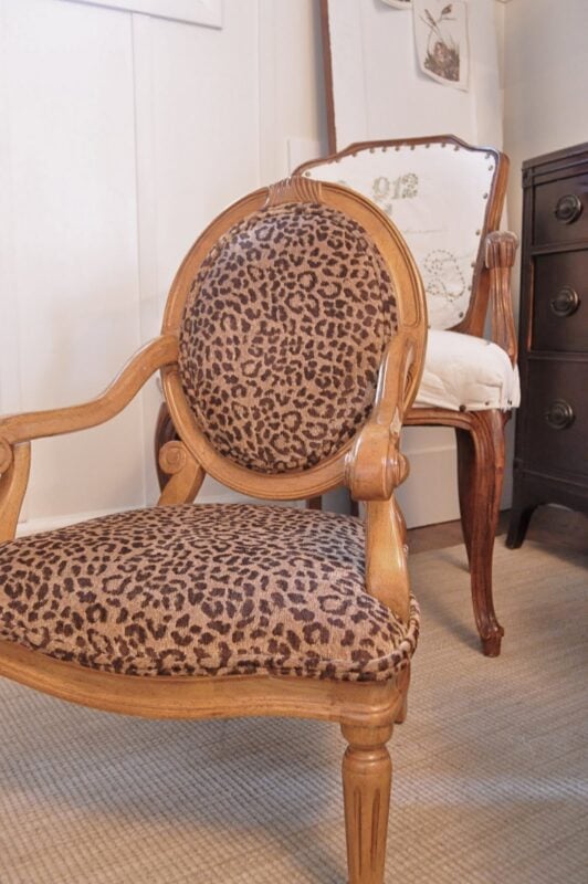 A wooden armchair with leopard print upholstery is in the foreground, while a reupholstered striped chair before and after transformation and a dark dresser are in the background.